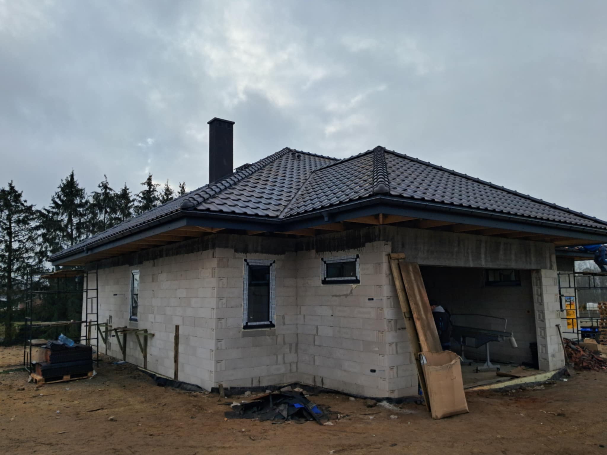 Nowy, grafitowy dach z kominem na surowym budynku z bloczków, z widocznymi rusztowaniami i materiałami budowlanymi. Pochmurne niebo w tle.