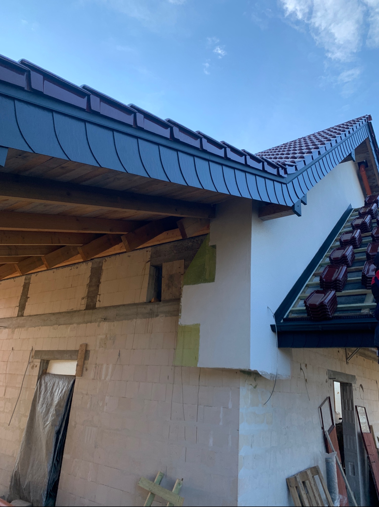 Widok na budynek w trakcie krycia dachu z użyciem dachówki ceramicznej. Część dachu pokryta dachówką karpiówką. Widoczne elementy konstrukcji dachu.