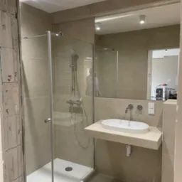 Beżowa łazienka w trakcie remontu: kabina prysznicowa ze szklanymi drzwiami, umywalka nablatowa na kamiennym blacie, lustro, widoczna surowa ściana z cegły