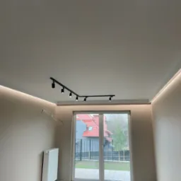 Jasne wnętrze z oświetleniem LED w listwach przysufitowych i reflektorami na szynie, widoczne okno balkonowe i instalacja elektryczna przed wykończeniem.