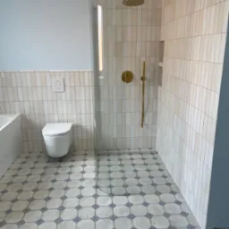 Wykończona łazienka z kabiną prysznicową typu walk-in, złotą armaturą i wzorzystą podłogą. Ściany wyłożone pionowymi, beżowymi płytkami. Widoczna toaleta i fragment wanny.