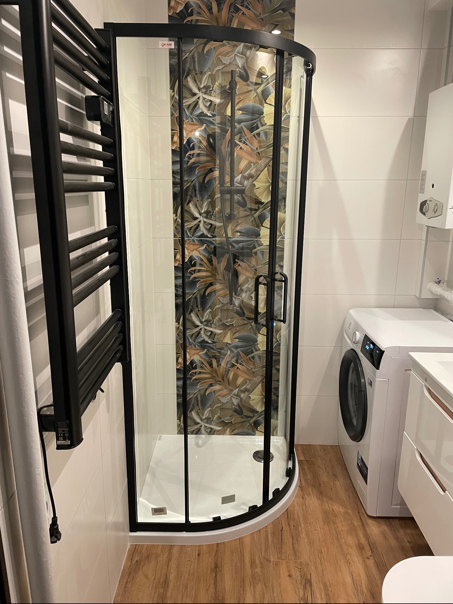 Nowoczesna kabina prysznicowa z czarną ramą i wzorzystą ścianą w tle, obok pralka i grzejnik drabinkowy w łazience z drewnopodobną podłogą.