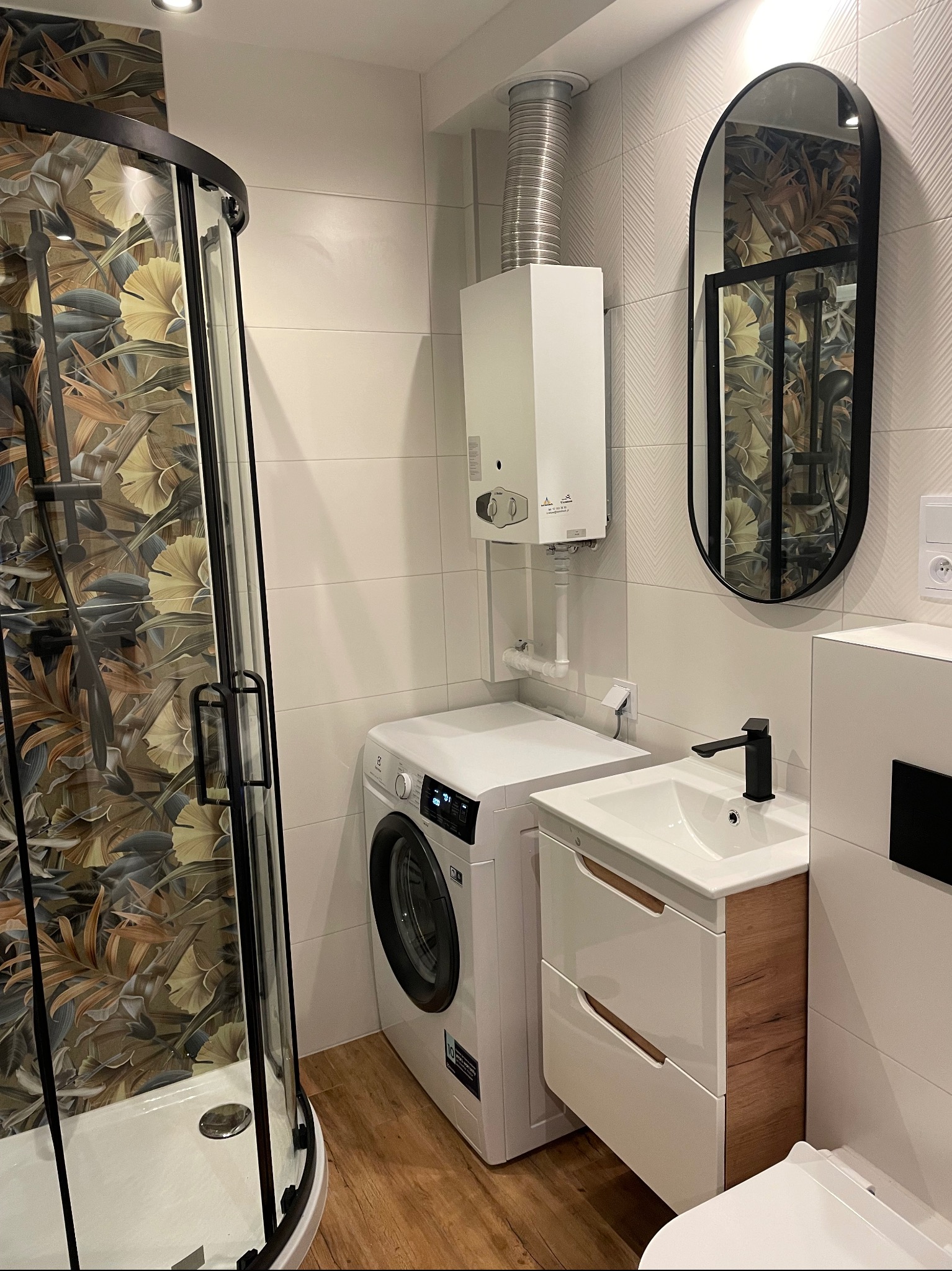 Nowoczesna łazienka z kabiną prysznicową w tropikalne wzory, pralką, umywalką z szafką w kolorze drewna i owalnym lustrem w czarnej ramie.