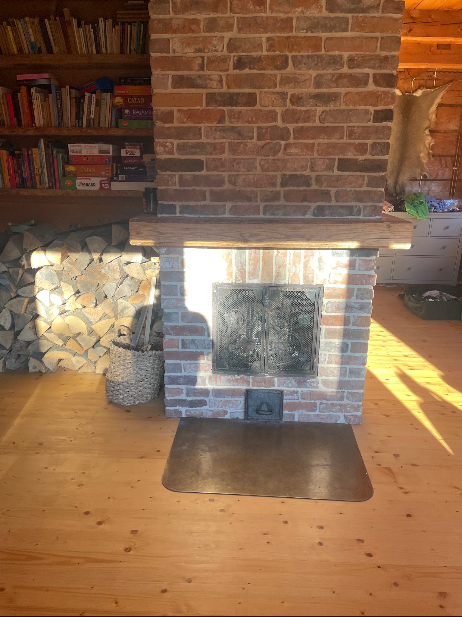 Ceglany kominek z drewnianą półką, metalowymi drzwiczkami z ornamentem i metalową płytą podłogową, obok kosz z drewnem, drewniana podłoga, regał z książkami w tle.
