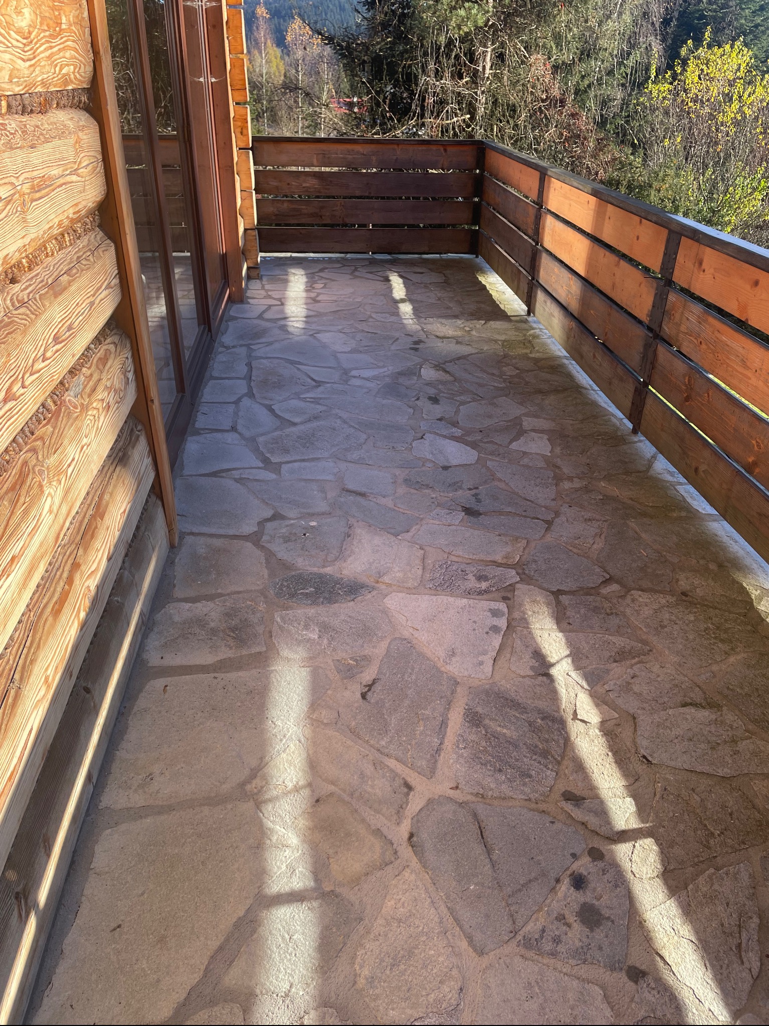 Balkon z drewnianą balustradą i kamienną posadzką, widok na zalesione wzgórze w słoneczny dzień.