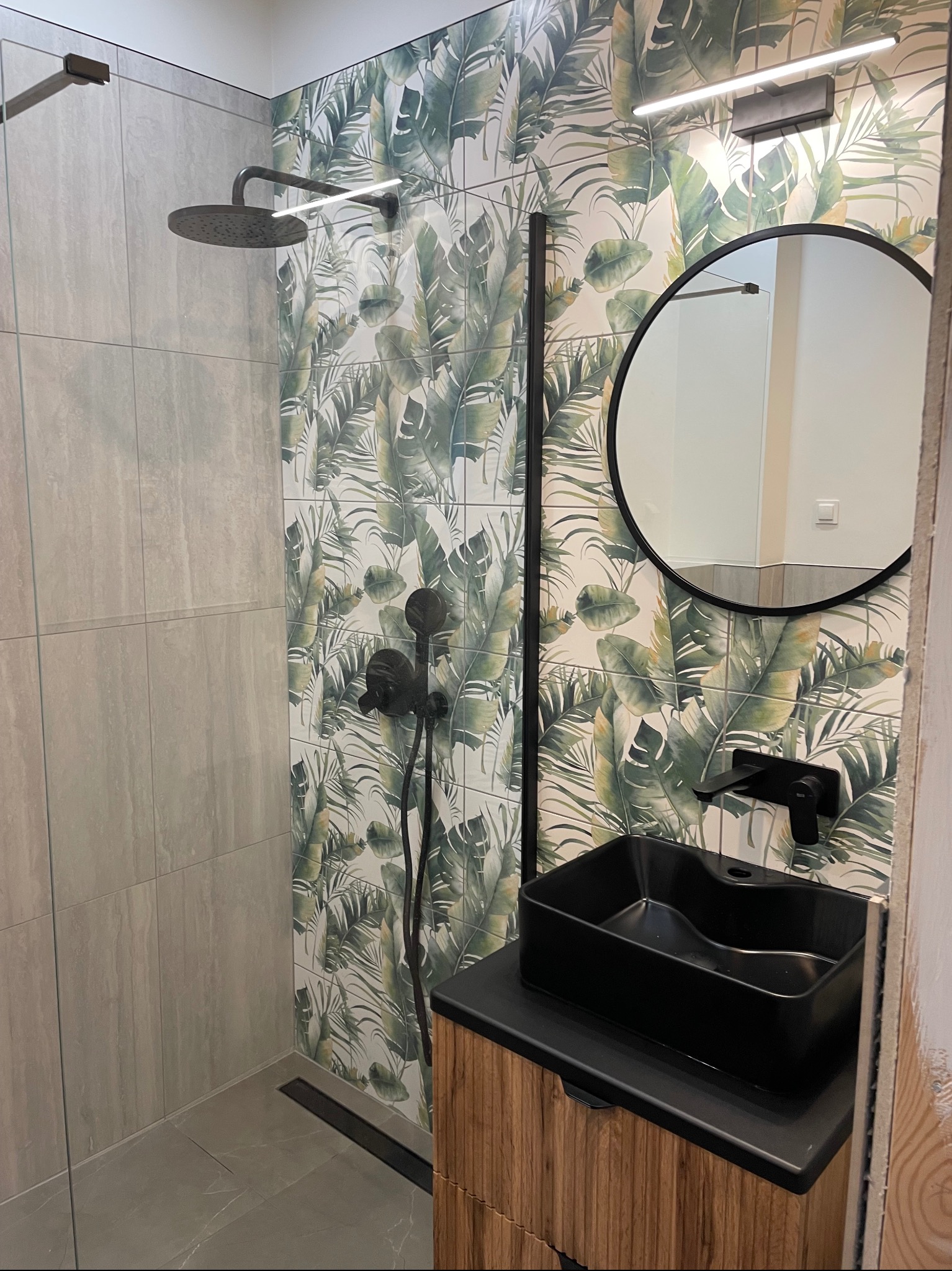 Nowoczesna łazienka z czarną armaturą, kabiną prysznicową walk-in i płytkami w tropikalne liście, okrągłe lustro z czarną ramą nad umywalką.