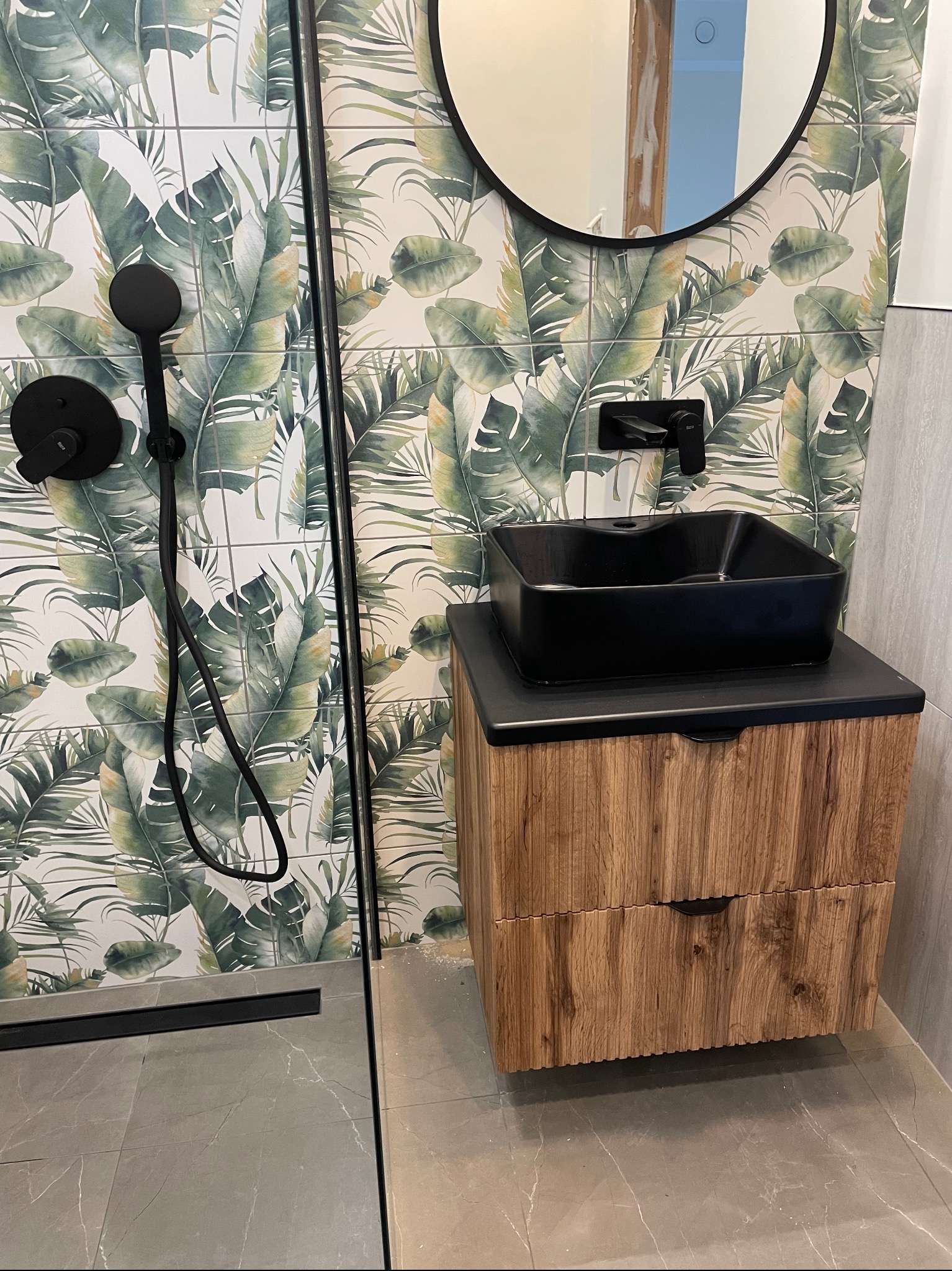 Nowoczesna łazienka z czarną, kwadratową umywalką nablatową na drewnianej szafce, tropikalne wzory na płytkach ściennych i czarna armatura.