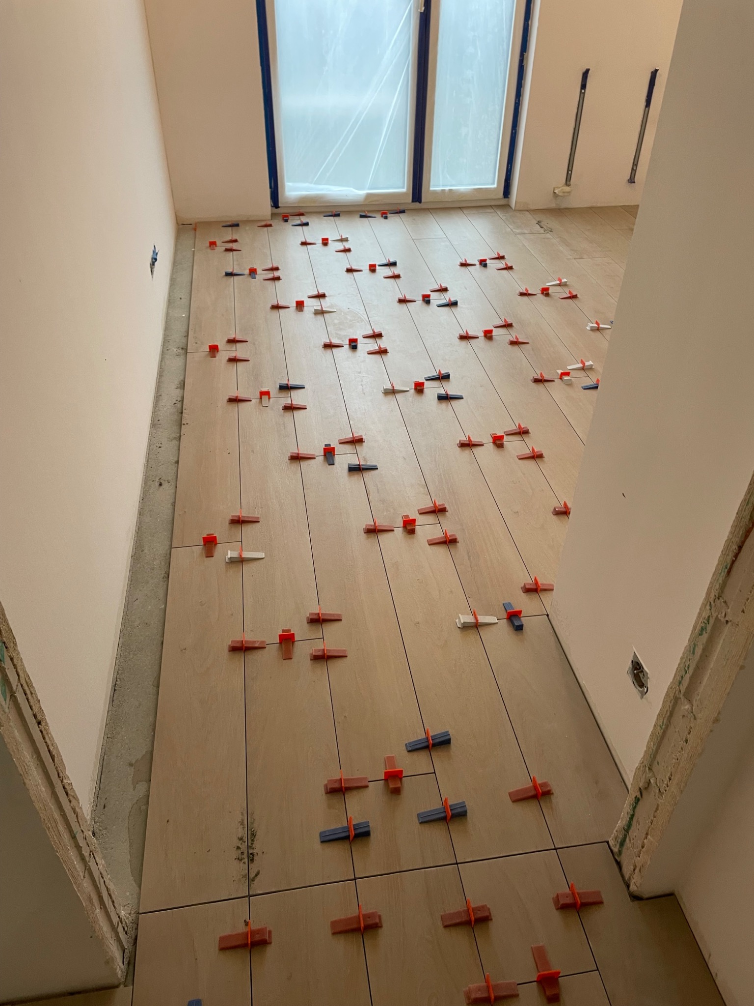 Układanie jasnych, podłużnych płytek podłogowych z wykorzystaniem systemu poziomowania płytek (klipsy i kliny) w trakcie prac wykończeniowych w pomieszczeniu z oknem.