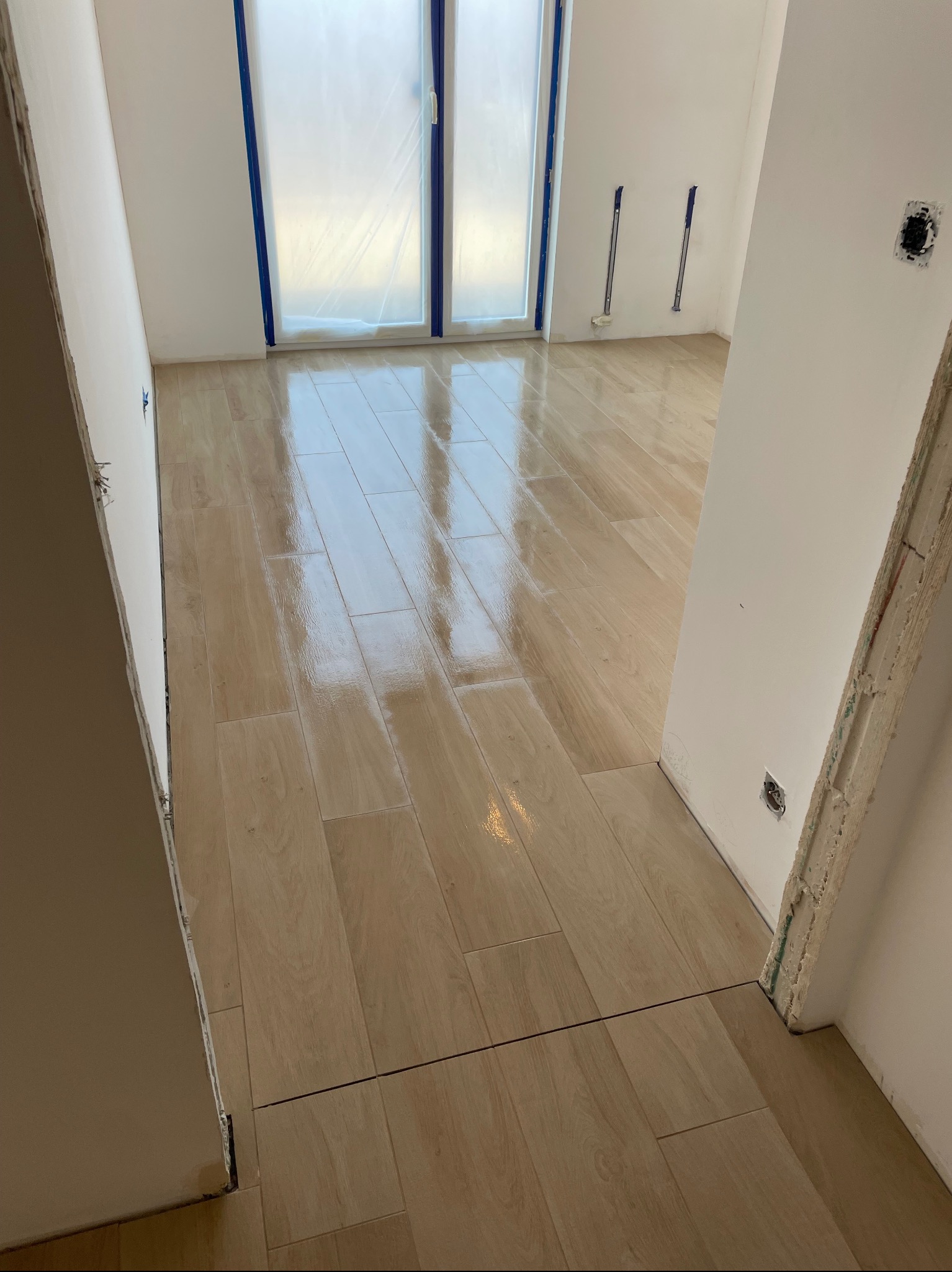Jasne, błyszczące płytki podłogowe imitujące drewno, ułożone w nowym mieszkaniu, widoczne przez otwarte drzwi, z zabezpieczonymi folią oknami i wystającymi kablami ze ściany.