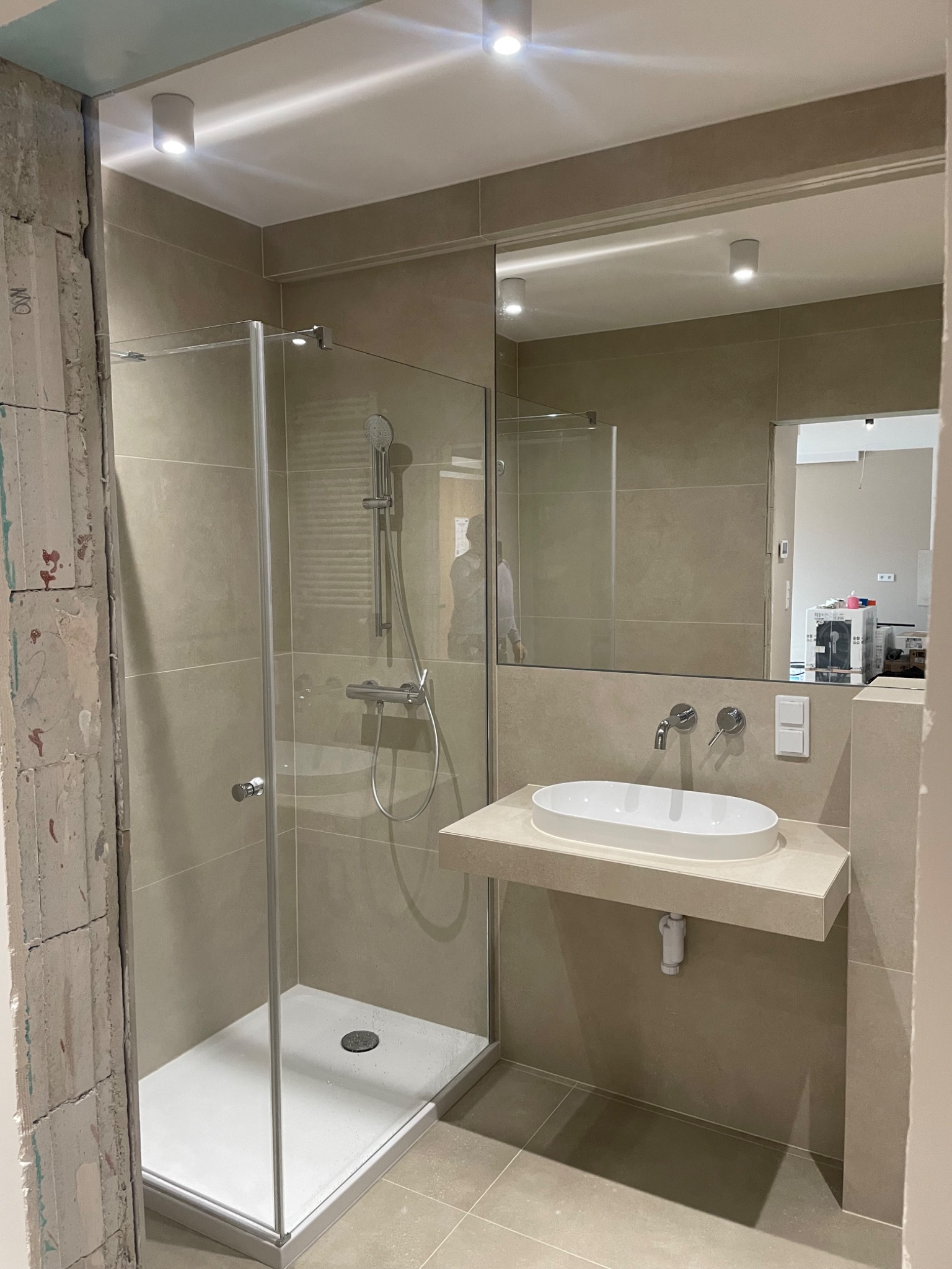 Beżowa łazienka w trakcie remontu: kabina prysznicowa ze szklanymi drzwiami, umywalka nablatowa na kamiennym blacie, lustro, widoczna surowa ściana z cegły