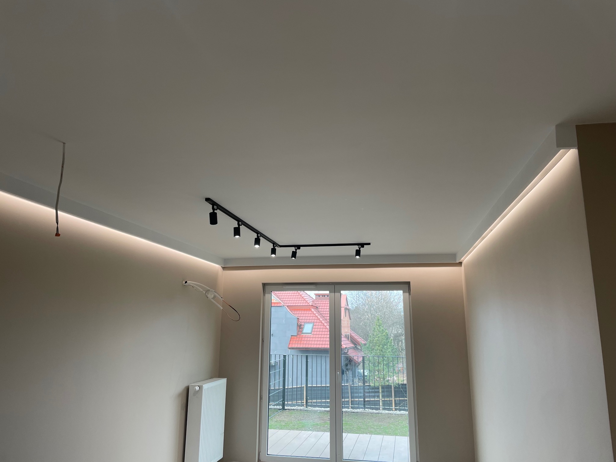 Jasne wnętrze z oświetleniem LED w listwach przysufitowych i reflektorami na szynie, widoczne okno balkonowe i instalacja elektryczna przed wykończeniem.