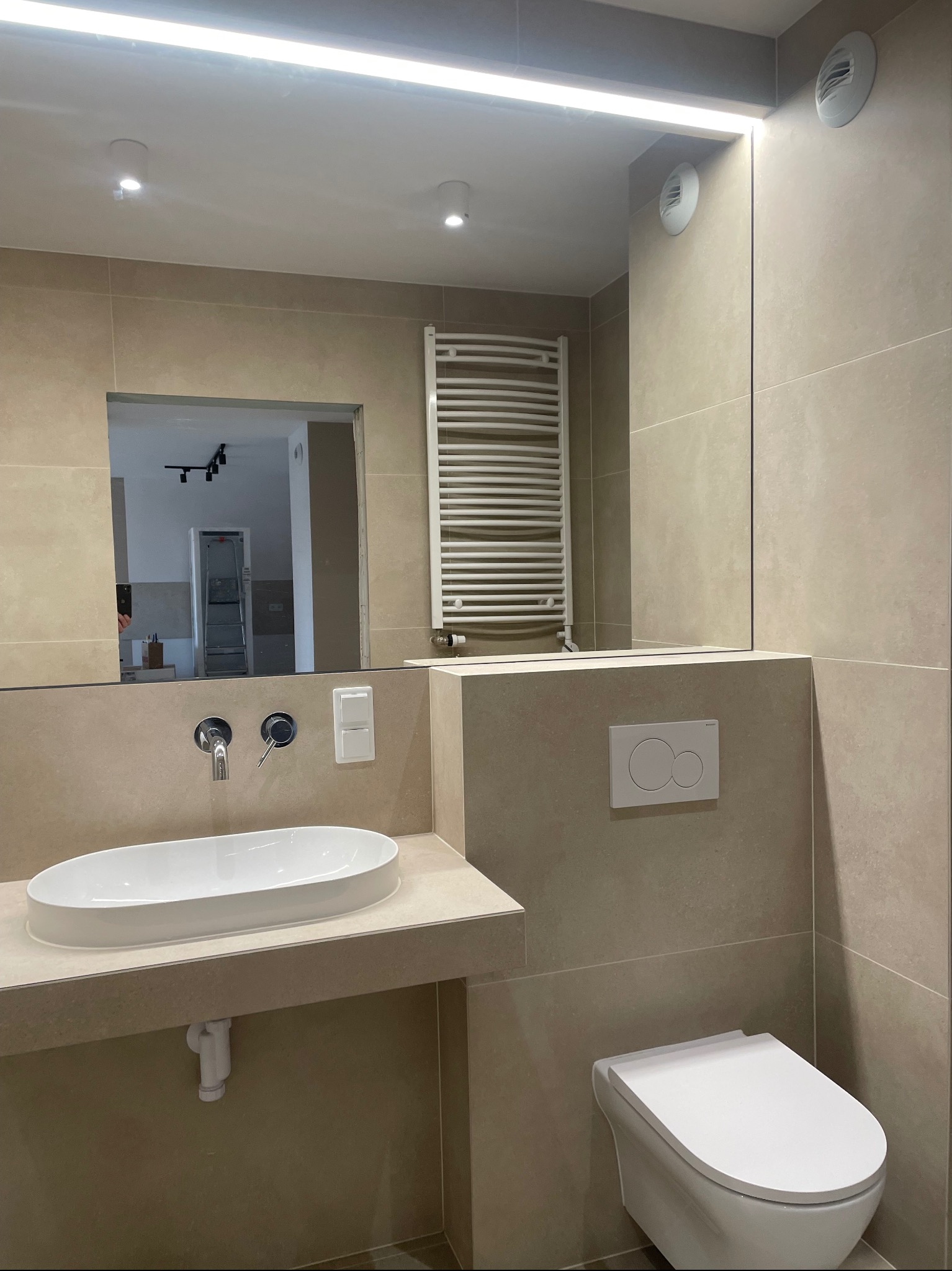 Beżowa łazienka z owalną umywalką nablatową, podświetlanym lustrem, grzejnikiem drabinkowym i wiszącą toaletą; w lustrze odbija się drabina budowlana.