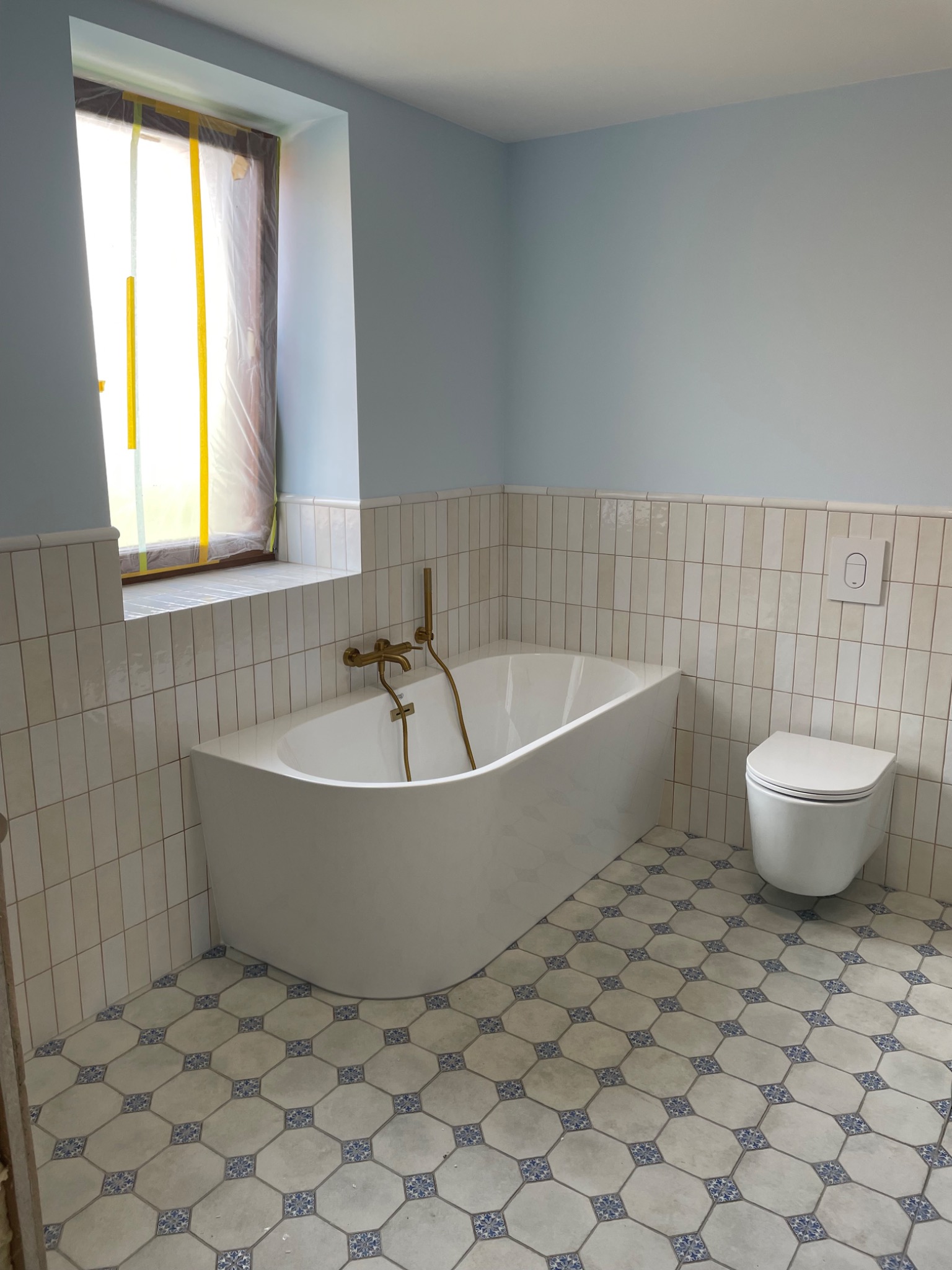 Wykończona łazienka w jasnych odcieniach: biała wanna z złotą armaturą, toaleta podwieszana i dekoracyjne płytki podłogowe. Okno zabezpieczone folią.