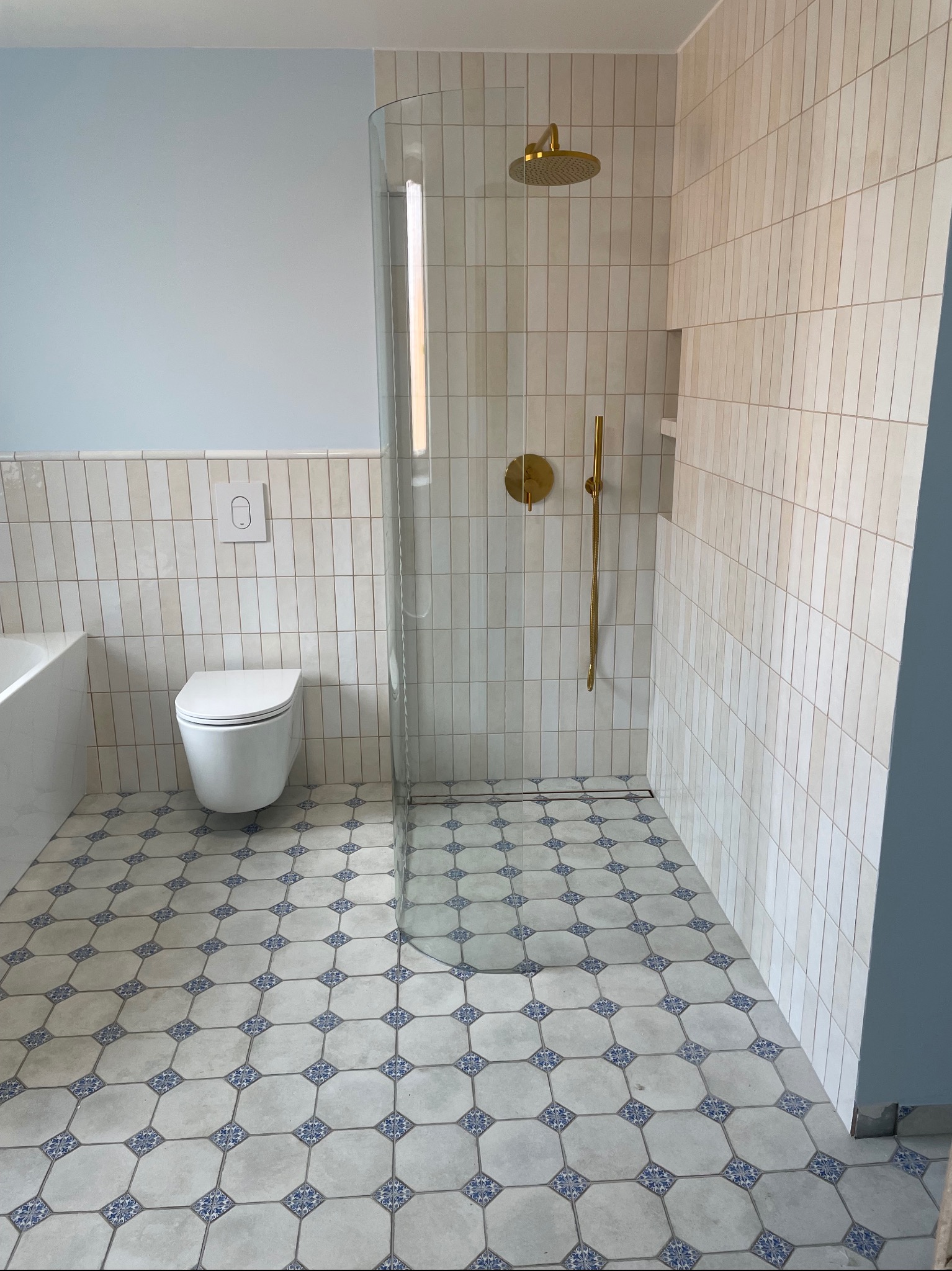 Wykończona łazienka z kabiną prysznicową typu walk-in, złotą armaturą i wzorzystą podłogą. Ściany wyłożone pionowymi, beżowymi płytkami. Widoczna toaleta i fragment wanny.