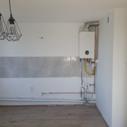 Grass and Garden Robert Zagrajek - Wnętrze kuchni w trakcie remontu: widoczne fragmenty płytek, rury, nowy piec gazowy Junkers oraz trzy lampy wiszące w stylu industrialnym.