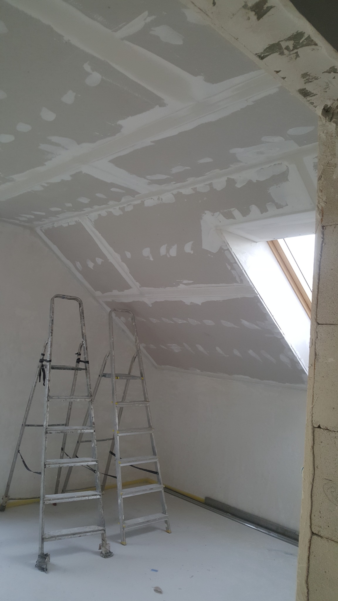 Sufit poddasza z zamontowaną płytą gipsowo-kartonową, widoczne łączenia wymagające szpachlowania, dwie aluminiowe drabiny ustawione w pomieszczeniu, okno dachowe w skosie.