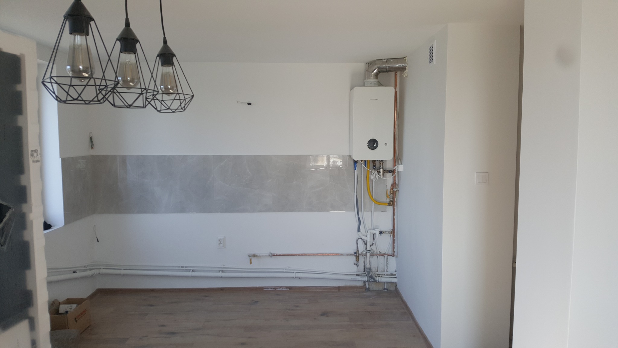 Wnętrze kuchni w trakcie remontu: widoczne fragmenty płytek, rury, nowy piec gazowy Junkers oraz trzy lampy wiszące w stylu industrialnym.