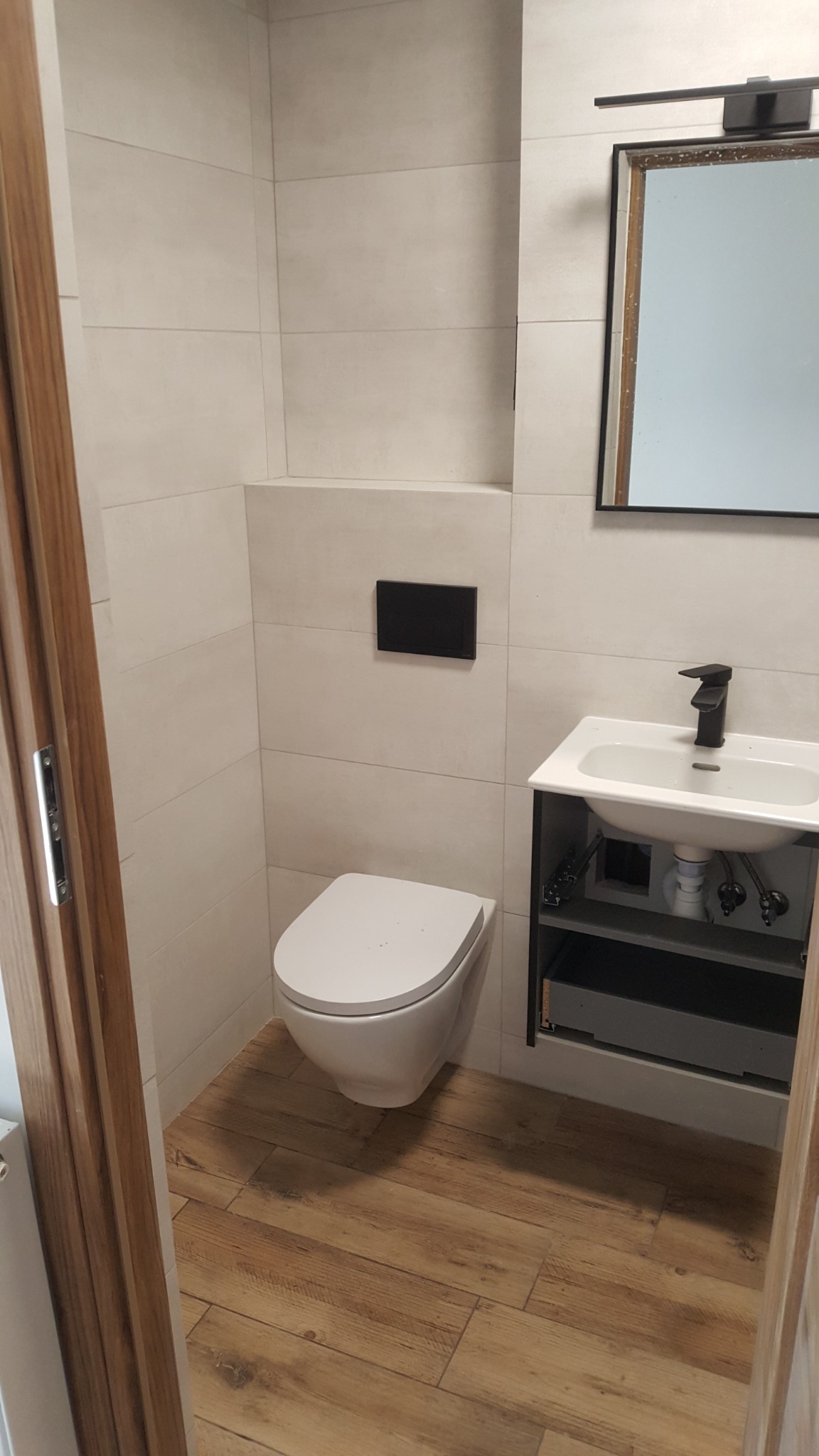 Nowoczesna łazienka z podwieszaną toaletą, umywalką nablatową z czarną armaturą i drewnopodobną podłogą, wykończona jasnymi płytkami.