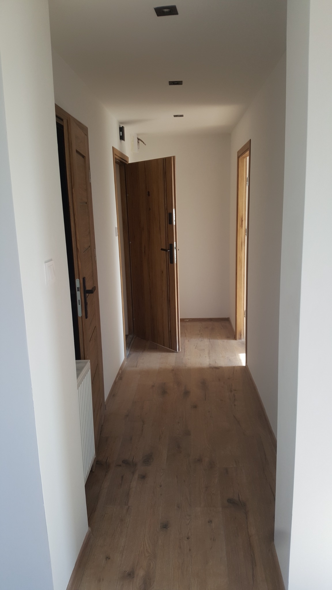 Korytarz po remoncie z drewnopodobnymi panelami podłogowymi, białymi ścianami i dwiema drewnianymi ościeżnicami drzwiowymi, z których jedne drzwi wejściowe są otwarte.