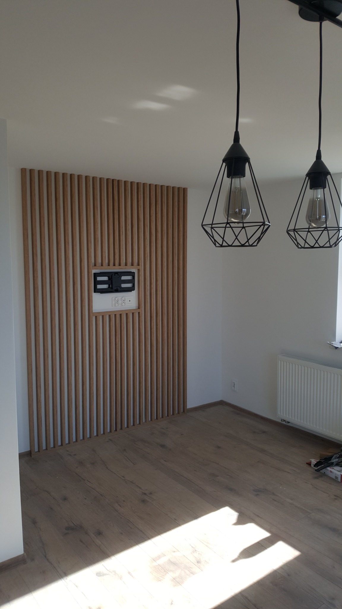 Drewniana ściana z listew z wnęką na telewizor i gniazdkami elektrycznymi, podłoga z paneli i wiszące lampy w pomieszczeniu w Żyrardowie.