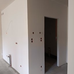 LUK-BUD Łukasz Słysz - Wnętrze w trakcie wykończenia: świeżo pomalowane ściany na biało, widoczne instalacje elektryczne i hydrauliczne, surowa podłoga, otwór drzwiowy. Minimalistyczny, surowy stan.