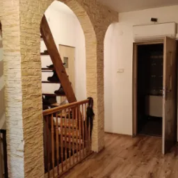 Aranżacja wnętrza z kamiennymi łukami i drewnianą podłogą. Widoczne schody z obuwiem i otwarte drzwi do pokoju. Klimatyczne oświetlenie.