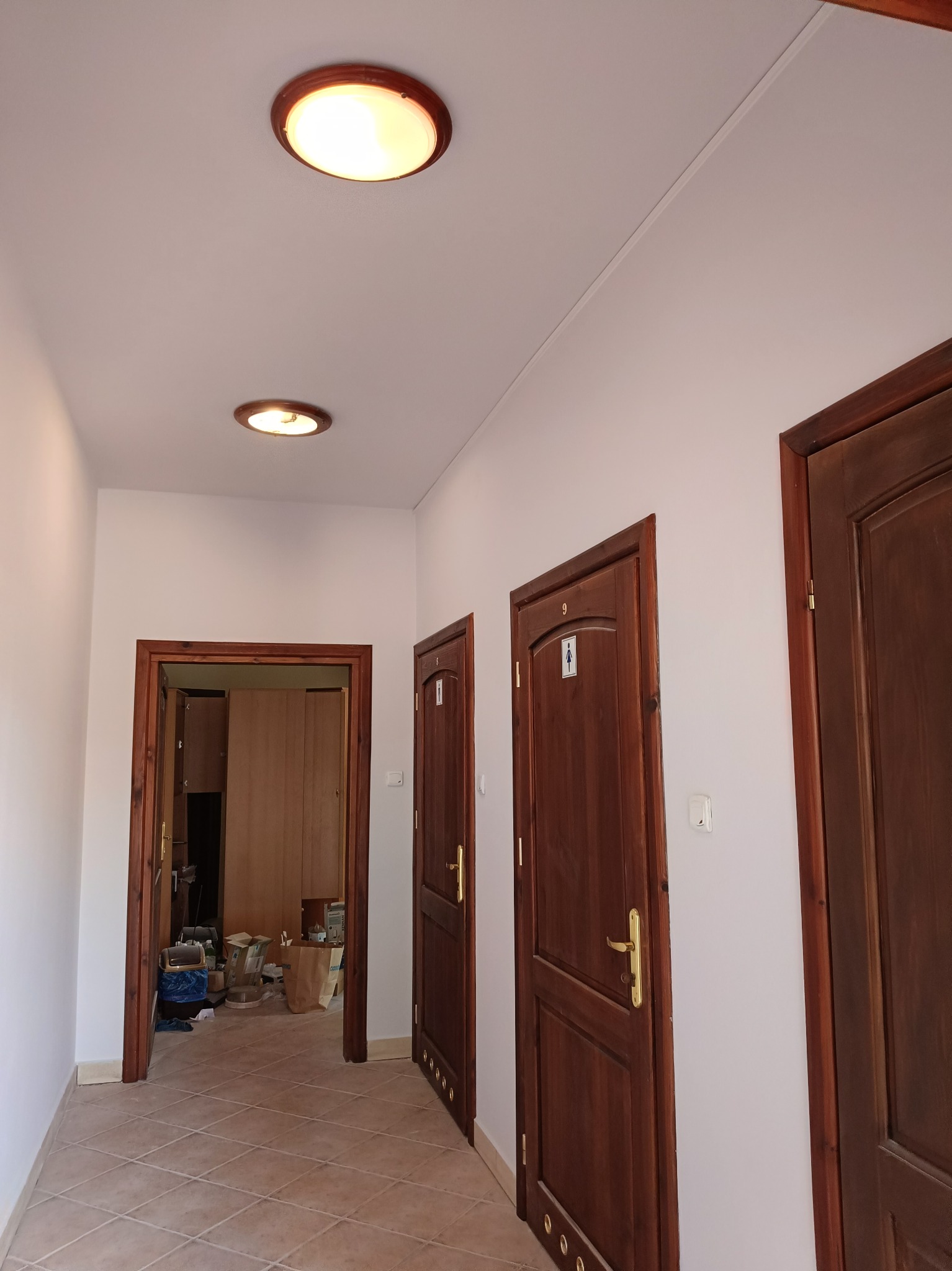 Korytarz z trzema drewnianymi drzwiami, w tym jedne otwarte ukazujące wnętrze. Na suficie dwa okrągłe plafony. Podłoga wyłożona płytkami. Świeżo pomalowane ściany.
