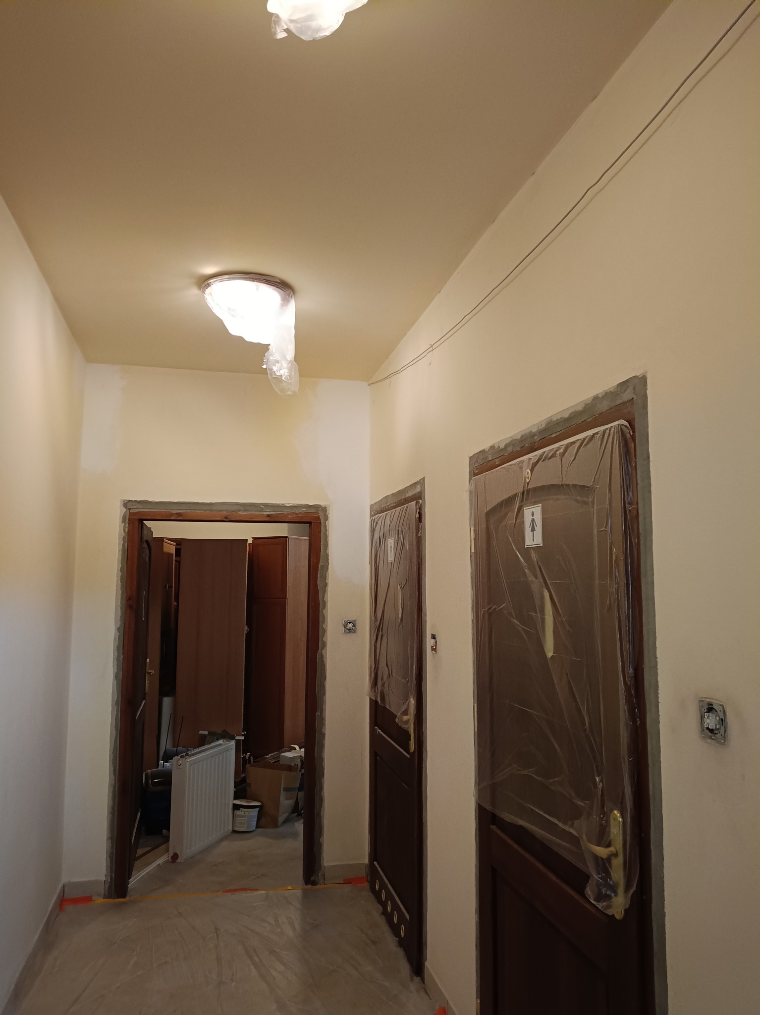 Korytarz w trakcie malowania ścian, zabezpieczone drzwi folią, widoczne niedomalowane fragmenty ścian. Grzejnik i inne przedmioty w tle sugerują trwający remont.