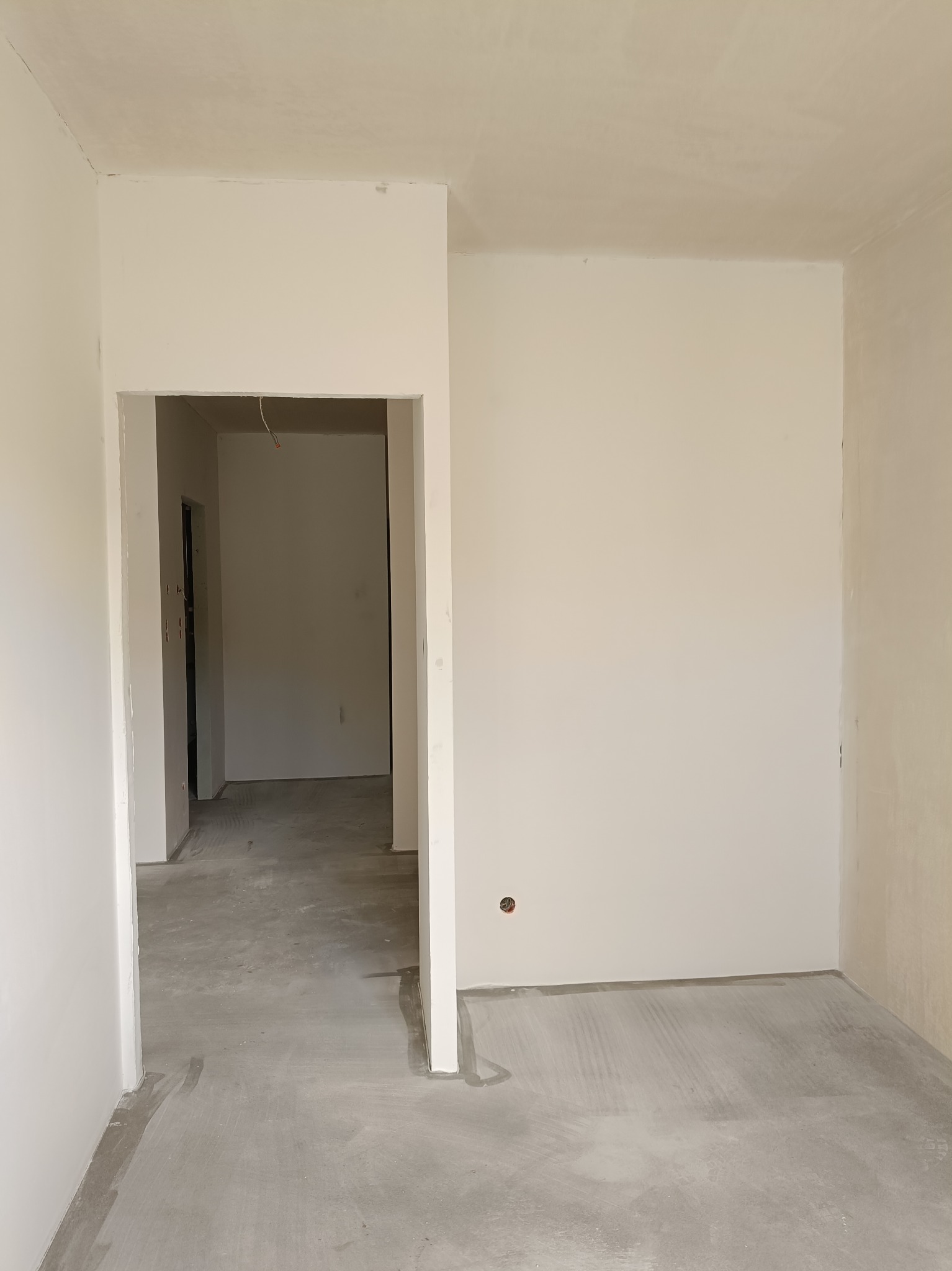 Wnętrze w trakcie wykończenia: białe ściany po szpachlowaniu, surowa podłoga i widoczne przejście do kolejnego pomieszczenia z elementami instalacji elektrycznej.