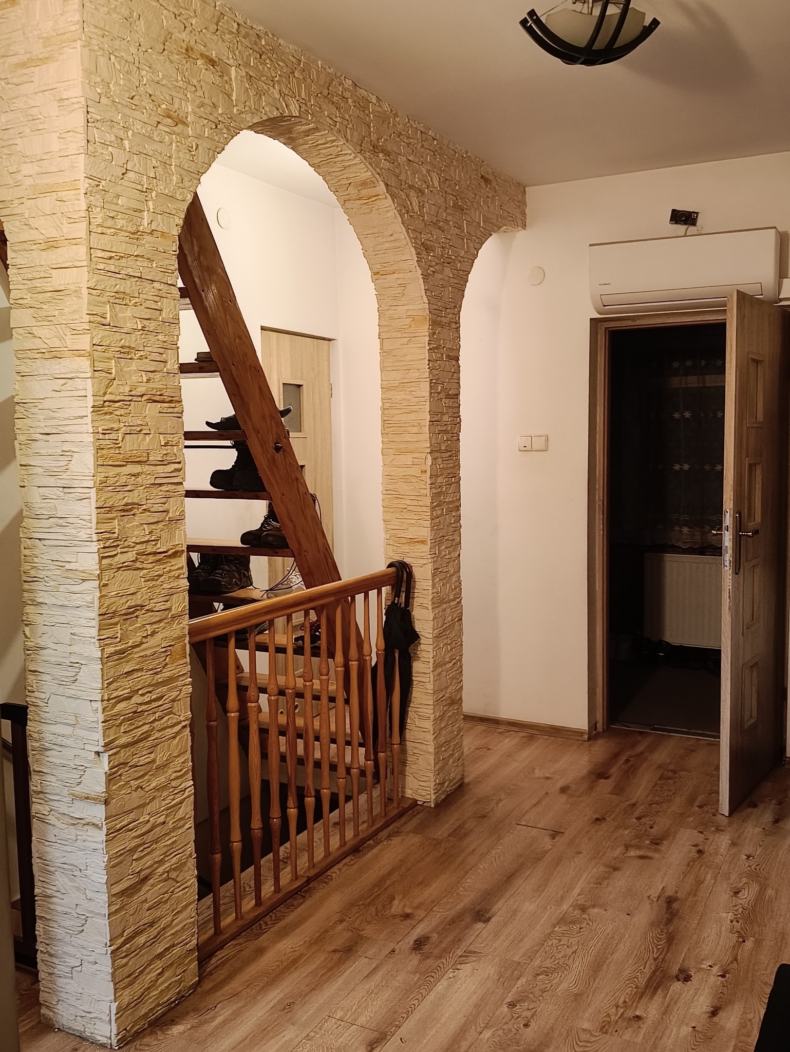 Aranżacja wnętrza z kamiennymi łukami i drewnianą podłogą. Widoczne schody z obuwiem i otwarte drzwi do pokoju. Klimatyczne oświetlenie.
