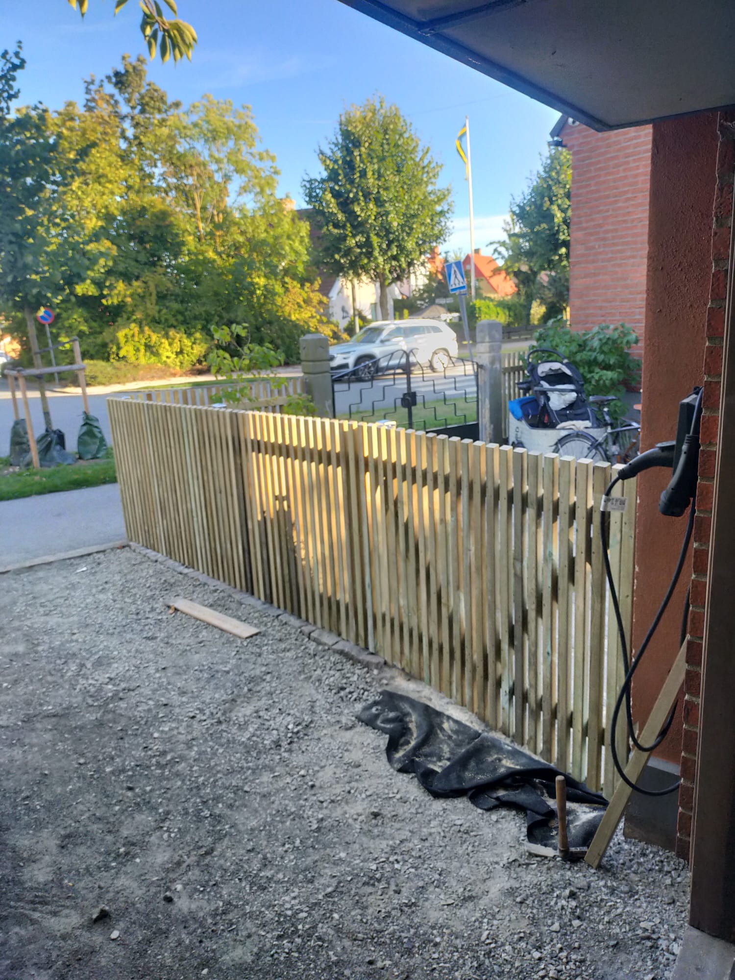 Nowe ogrodzenie z pionowych desek przed posesją, widoczne narzędzia i czarna plandeka na żwirze.