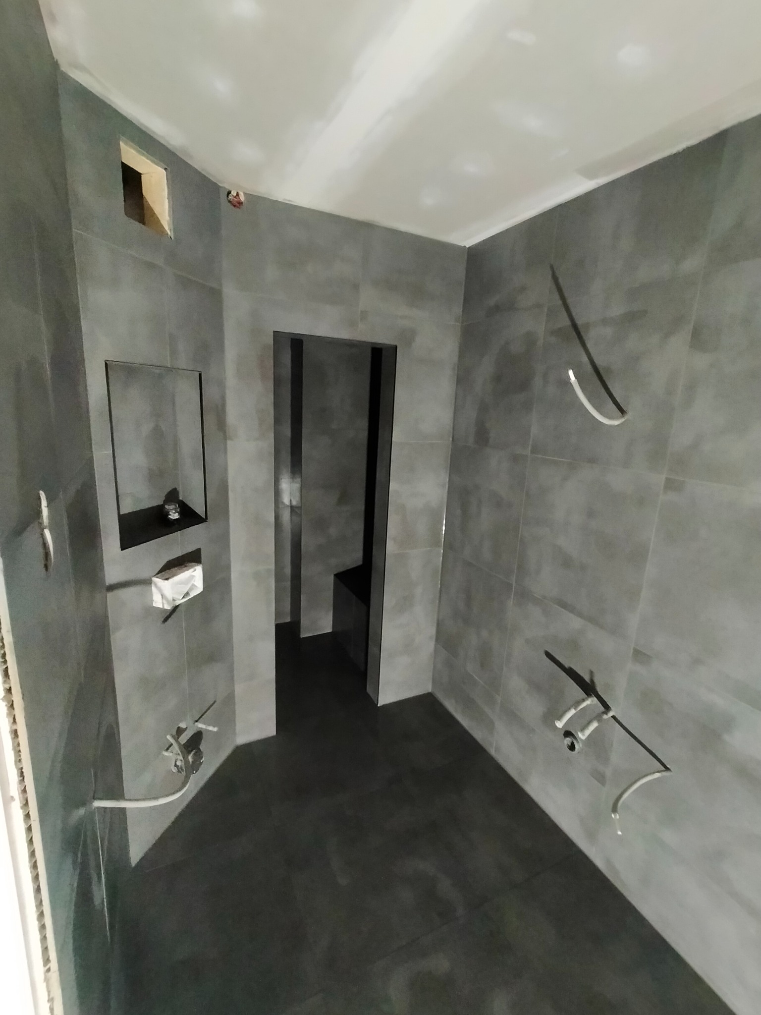 Wnętrze łazienki w trakcie prac wykończeniowych: szare płytki na ścianach i podłodze, widoczne wystające rury instalacyjne, wnęka w ścianie z półką, surowy sufit.