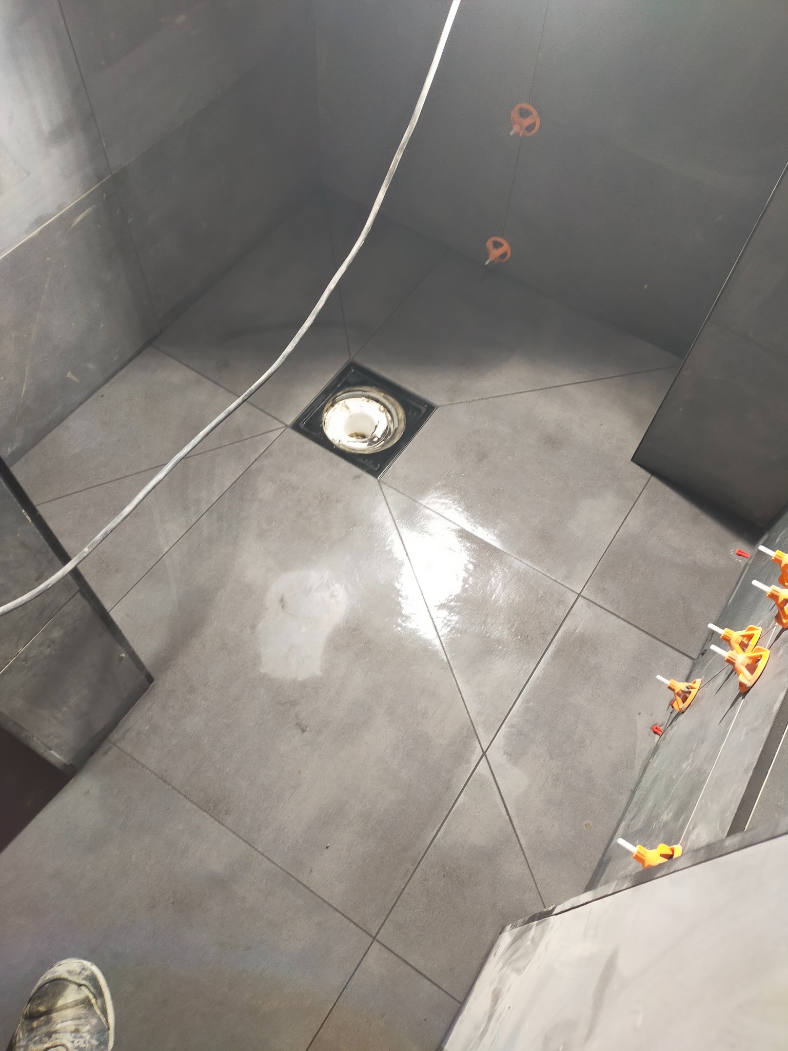 Szare płytki ułożone w romby na podłodze i ścianach prysznica, z widocznym odpływem liniowym i systemem poziomowania płytek, w trakcie prac wykończeniowych.