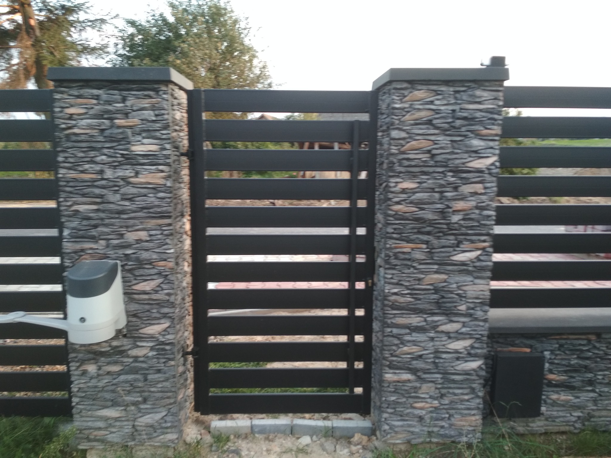 Nowoczesna brama wjazdowa z poziomych, czarnych profili, zamontowana pomiędzy kamiennymi słupkami z automatyką do otwierania, widoczna z zewnątrz posesji.