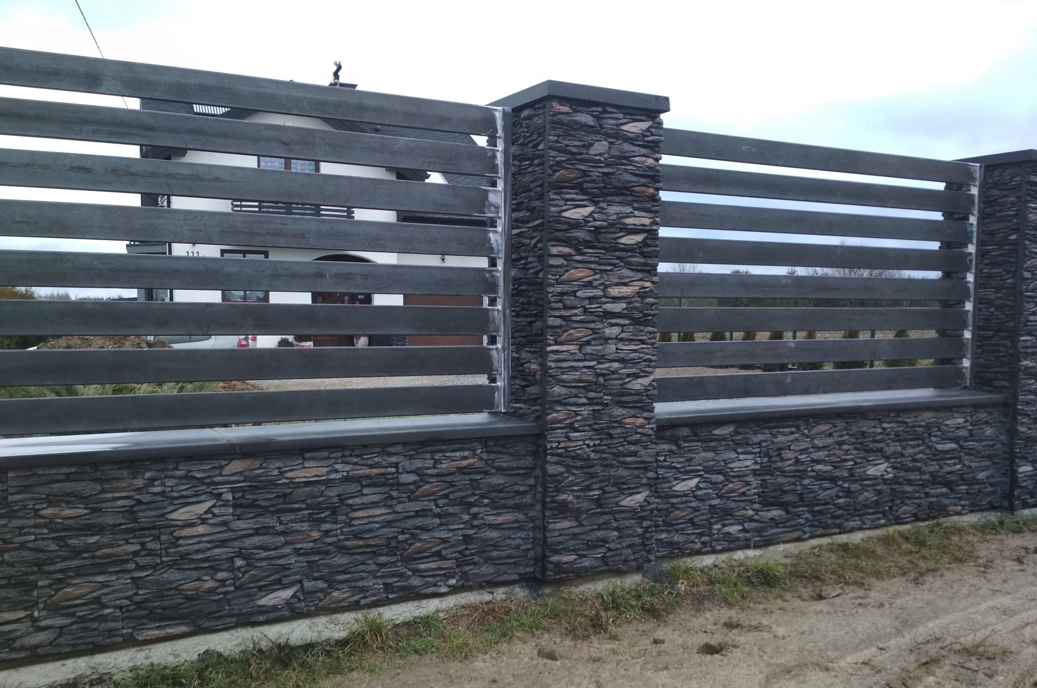 Nowoczesne ogrodzenie z poziomych, szarych desek i kamiennych słupków, widok z zewnątrz posesji na tle domu.