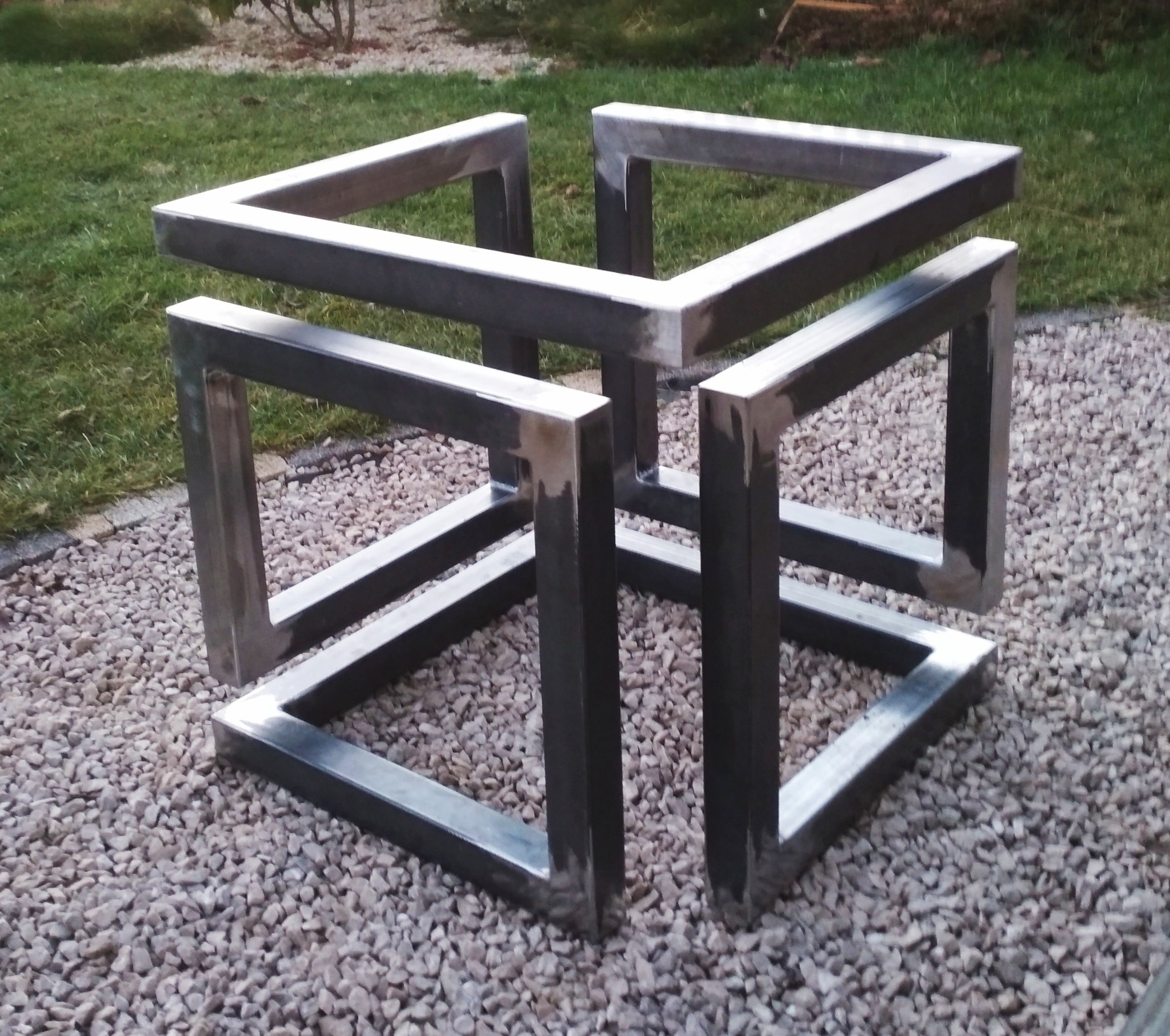 Metalowa konstrukcja przestrzenna w kształcie sześcianu, wykonana z profili, ustawiona na żwirowym podłożu z trawnikiem w tle.