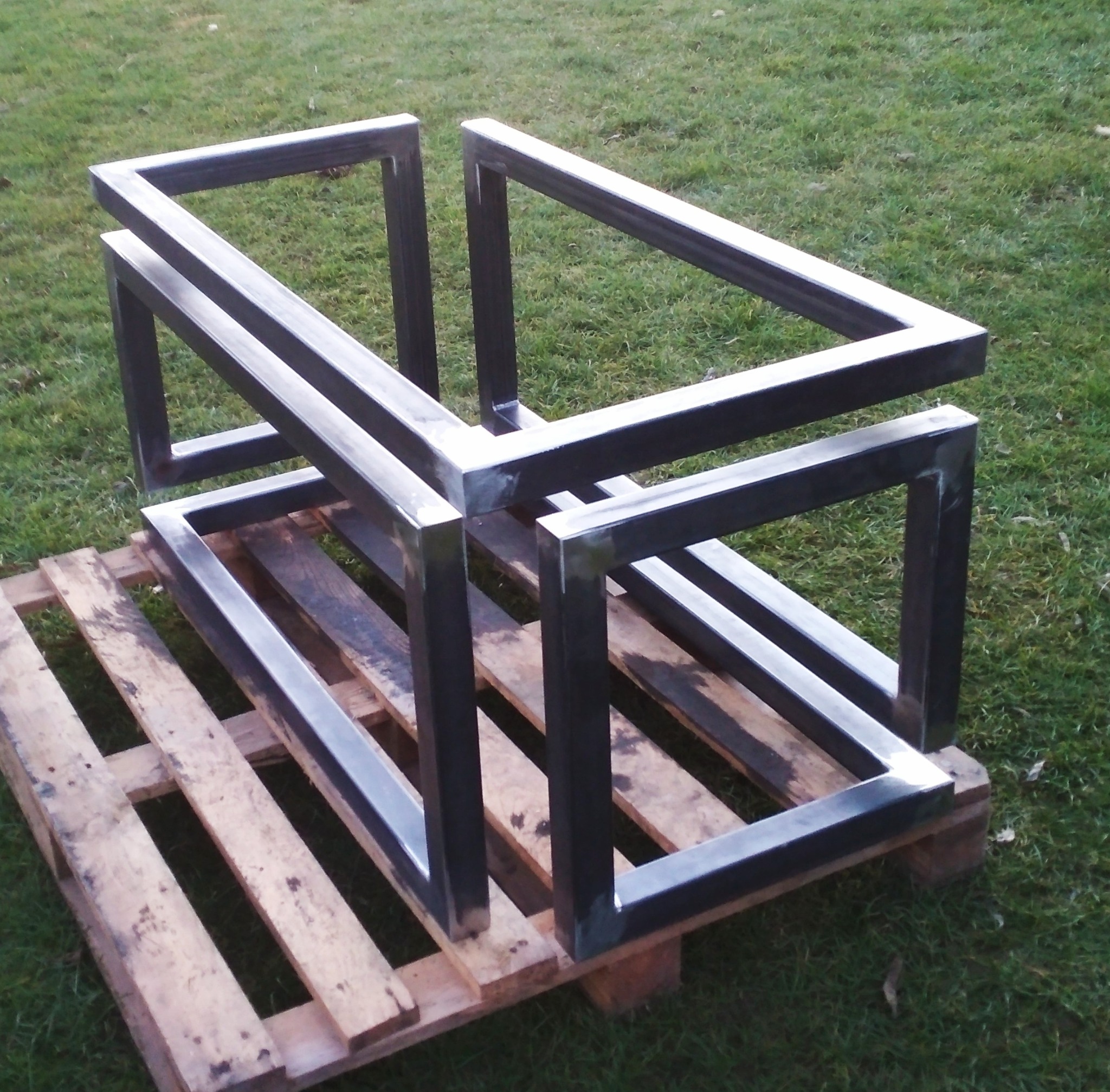 Surowa, metalowa konstrukcja z profili kwadratowych, spawana, ustawiona na drewnianej palecie na trawie.