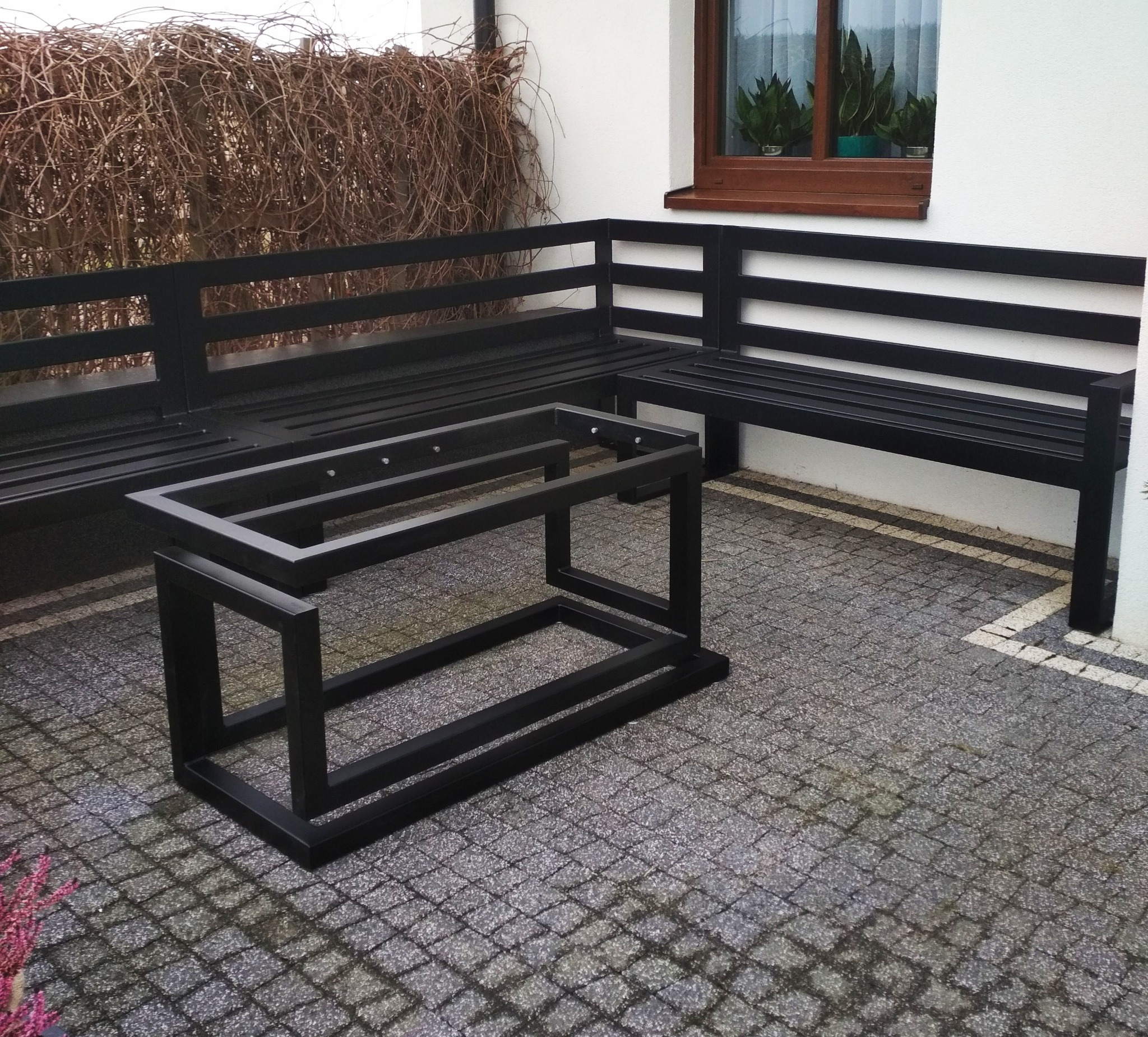 Czarny metalowy narożnik z ławkami i stolikiem o minimalistycznej konstrukcji na tle jasnej ściany i brukowanej nawierzchni.