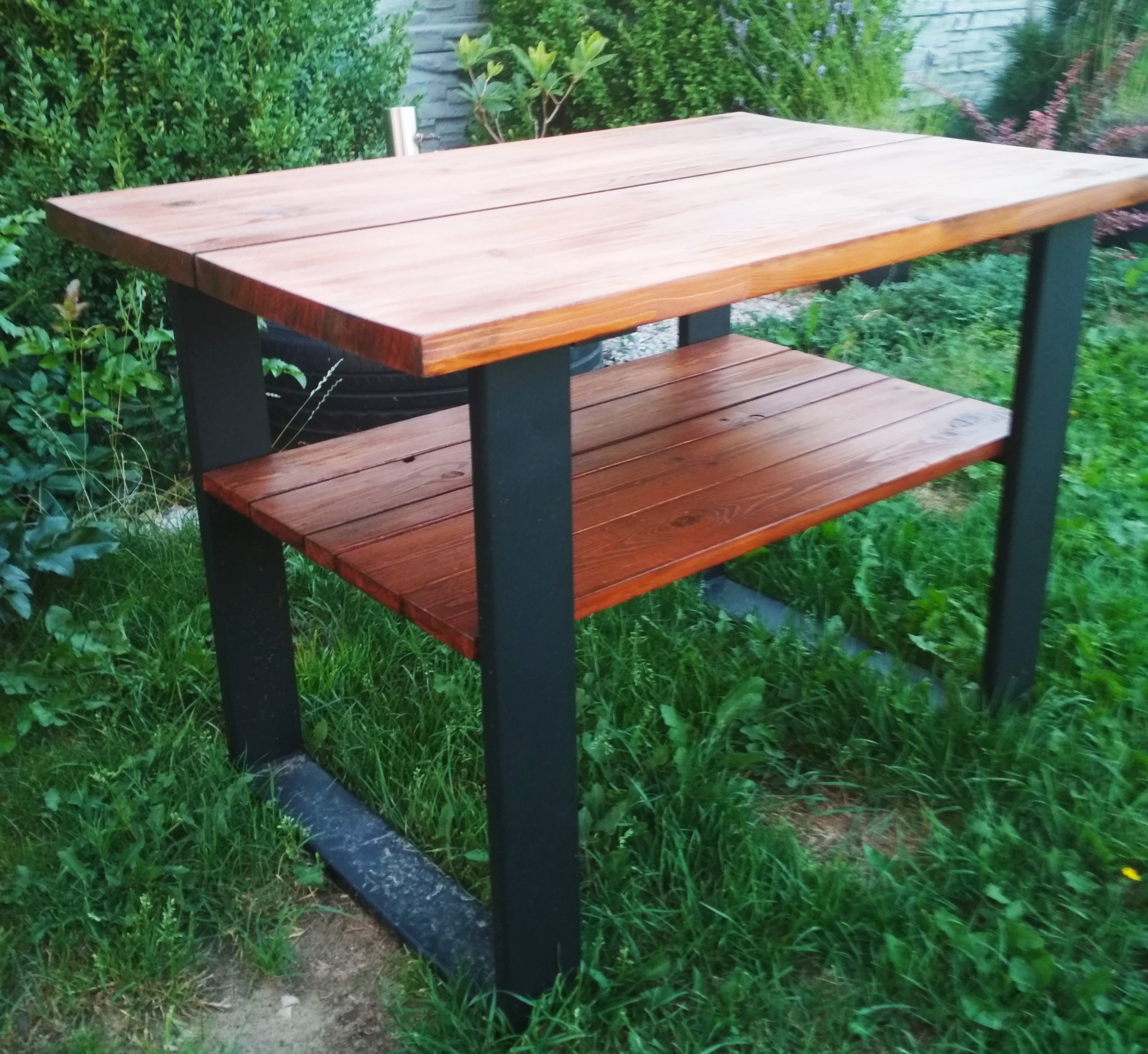 Drewniany stół ogrodowy z metalową konstrukcją, dwie poziome powierzchnie, ustawiony na trawie w otoczeniu zieleni.