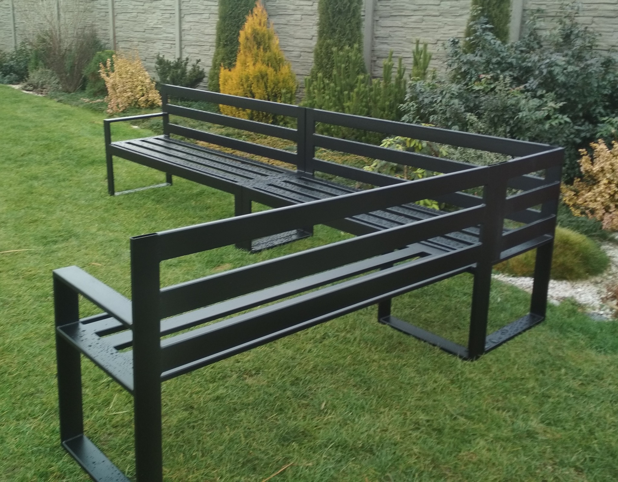 Nowoczesna, minimalistyczna pergola metalowa w kolorze grafitowym, z elementami siedziska, umieszczona na trawniku w ogrodzie z tujami i kamiennym ogrodzeniem w tle.