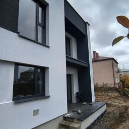 MixBud - Dwukondygnacyjny dom z białą elewacją i czarnymi akcentami architektonicznymi, widok z boku na niezagospodarowany teren działki i sąsiedni budynek w tle.