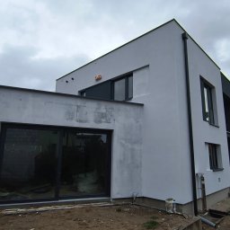 MixBud - Nowoczesny dom z elewacją w trakcie wykończenia, widoczne ślady po gładzi na części ściany, duże okna z czarnymi ramami, teren wokół domu częściowo przygotowany pod trawnik.