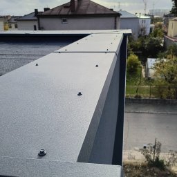 MixBud - Szczegółowe ujęcie szarej, metalowej obudowy balkonu z widocznymi śrubami mocującymi, na tle zabudowy miejskiej i pochmurnego nieba.