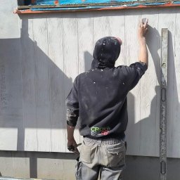 MixBud - Pracownik w bluzie z kapturem, widziany od tyłu, wygładza szpachlą ścianę budynku z oknem zabezpieczonym folią, obok stoi poziomica.