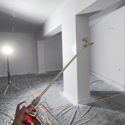 MixBud - Malowanie ścian w pomieszczeniu biurowym za pomocą pistoletu natryskowego, widoczne zabezpieczenia podłogi folią i oświetlenie halogenowe.