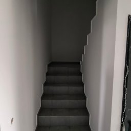 MixBud - Schody wewnętrzne wyłożone ciemnymi płytkami, prowadzące w górę, ściany pomalowane na jasny odcień szarości, widoczny fragment uszkodzonej ściany i obudowy drzwi.