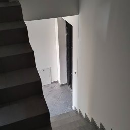 MixBud - Klatka schodowa z ciemnymi schodami wyłożonymi płytkami, prowadząca w dół do korytarza z szarą podłogą i białymi ścianami, widoczne drzwi wejściowe.