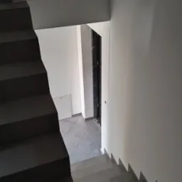 Klatka schodowa z ciemnymi schodami wyłożonymi płytkami, prowadząca w dół do korytarza z szarą podłogą i białymi ścianami, widoczne drzwi wejściowe.