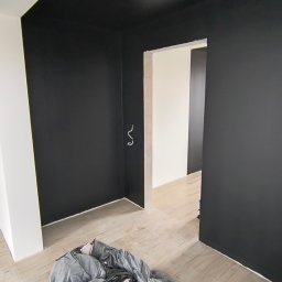 MixBud - Pomieszczenie w trakcie malowania: czarny sufit i ściany, zabezpieczona podłoga folią malarską, widoczne przewody elektryczne wystające ze ściany.