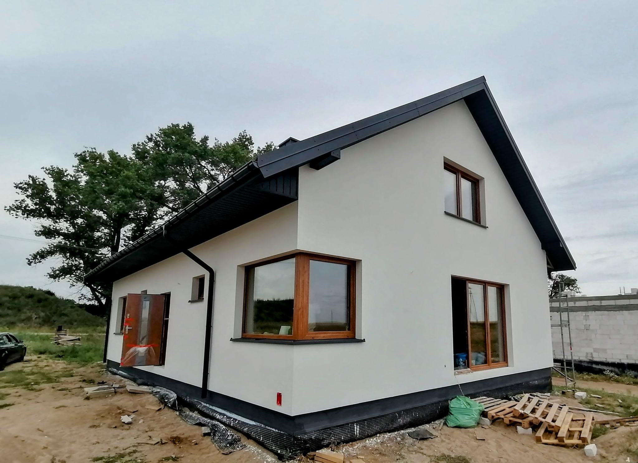 Nowoczesny, parterowy dom z białą elewacją i ciemnym dachem, widok z zewnątrz, teren wokół w trakcie zagospodarowania, widoczne palety i worki budowlane.